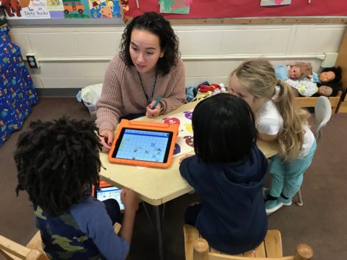 Photo of Sarah seated at a table with students looking at a tablet